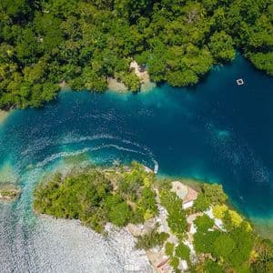 Luftaufnahme eines kleinen Bootes mit Kielwelle auf einer tiefblauen Lagune, umgeben von üppigem Grünwald.