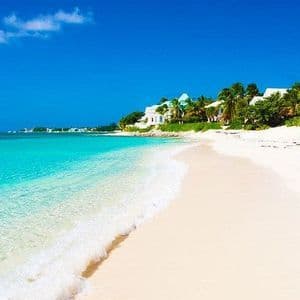 Ein weißer Sandstrand mit klarem, türkisfarbenem Wasser, das ans Ufer plätschert, gesäumt von Palmen und Villen unter einem strahlend blauen Himmel.