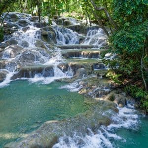 Ein terrassierter Wasserfall stürzt über Felsen in ein klares, türkisfarbenes Becken, umgeben von üppiger grüner Dschungelvegetation.