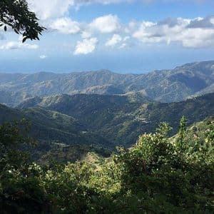 Ein malerischer Ausblick auf üppige, grüne Berge und Täler, die sich bis zum Meer erstrecken, unter einem Himmel mit flauschigen weißen Wolken.