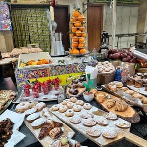 Una bancarella del mercato espone dolci freschi come ciambelle e cornetti, accanto a una pila di arance e uno spremiagrumi.