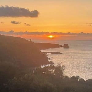 Un tramonto arancione sull'oceano accanto a una costa collinare con un piccolo molo e nuvole sparse.