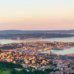Una vista dall'alto di una città costiera e del suo porto, illuminata dalla luce calda del tramonto, con colline che circondano la baia.
