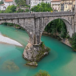 Un ponte storico in pietra si staglia su un fiume turchese, con un centro storico e un campanile visibili sulla riva opposta sotto un cielo nuvoloso.