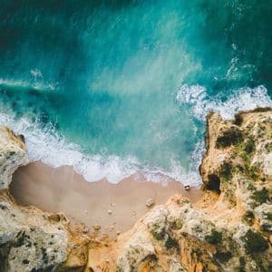 Vue aérienne de vagues turquoise de l'océan s'écrasant sur une plage de sable isolée, entourée de hautes falaises rocheuses.