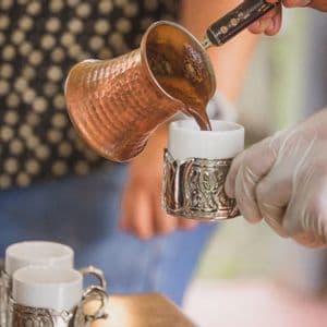 Una persona vierte café tradicional de una cafetera de cobre en una pequeña taza ornamentada sostenida por una mano enguantada.