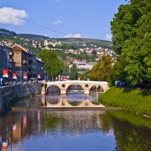 Un puente de piedra con múltiples arcos atraviesa un río que serpentea por una ciudad, con edificios en una orilla y árboles en la otra.