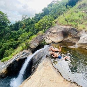 Un uomo e una donna si rilassano in una piscina naturale rocciosa poco profonda ai margini di una cascata, con vista su una fitta foresta verde.