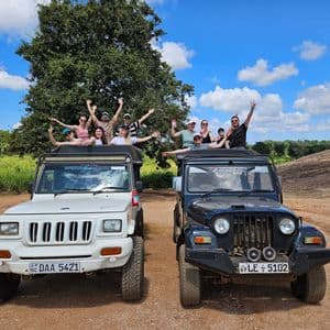 Un viaggio di gruppo WeRoad sorride e saluta dai tetti aperti di due jeep da safari parcheggiate su una strada sterrata sotto un cielo azzurro.