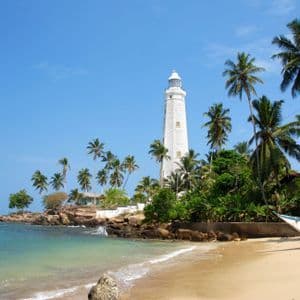 Un alto faro bianco si erge su una punta rocciosa circondata da palme su una spiaggia tropicale.
