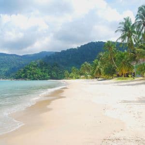 Una spiaggia di sabbia bianca con acqua turchese, fiancheggiata da palme di fronte a verdi colline lussureggianti sotto un cielo parzialmente nuvoloso.