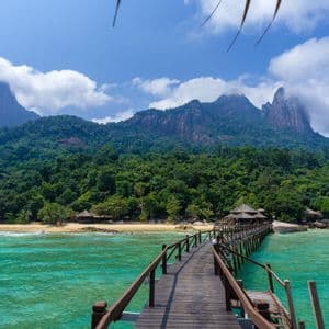 Un lungo molo di legno si estende su acque turchesi verso una spiaggia sabbiosa, con dietro montagne verdi e lussureggianti sotto un cielo azzurro con nuvole soffici.