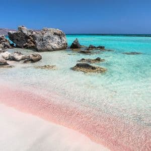 Sabbia rosa su una spiaggia bianca incontra acque turchesi cristalline con grandi rocce sotto un cielo blu.