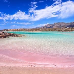 Una spiaggia di sabbia rosa incontra acque limpide e turchesi, con montagne in lontananza sotto un cielo azzurro e nuvole bianche.