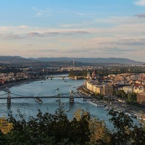 Una vista panoramica di una città costruita lungo un ampio fiume, con diversi ponti, barche e dolci colline in lontananza sotto un cielo tenue.