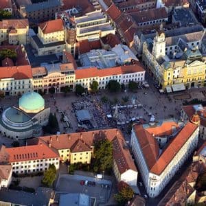 Una vista aerea di una piazza europea affollata, caratterizzata da una grande cupola verde e edifici con tetti tradizionali in tegole rosse.