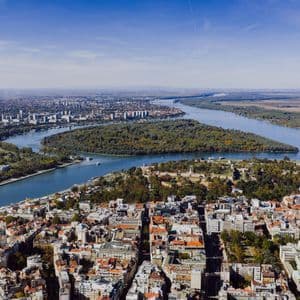 Vista aerea di una vasta città con un ampio fiume che si biforca, scorrendo attorno a una grande isola boscosa sotto un cielo azzurro.