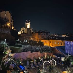 Una terrazza panoramica con lettini, luci decorative e statue di giraffa, con vista su un castello illuminato e sul paesaggio urbano notturno.
