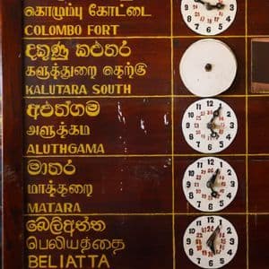 Un primo piano di un tabellone delle partenze ferroviarie in legno, che mostra destinazioni come Colombo Fort e Matara accanto a orologi analogici.