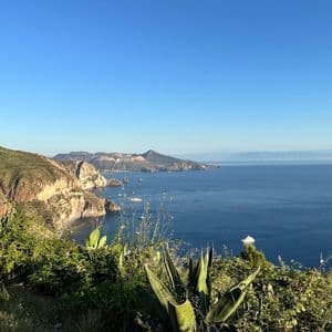 Un panorama da una collina verde che si affaccia su una costa rocciosa, un mare blu profondo con barche e isole vulcaniche all'orizzonte.