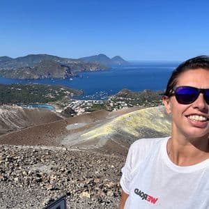 Una donna sorridente con una maglietta WeRoad e occhiali da sole si scatta un selfie da una cima vulcanica che si affaccia su una baia con isole.
