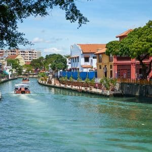 Due battelli turistici navigano un fiume costeggiato da edifici storici e colorati e alberi verdi lussureggianti sotto un cielo azzurro limpido.