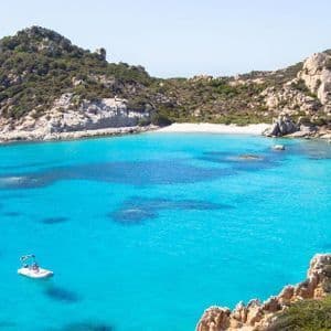 Numerose barche e barche a vela sono ormeggiate in una baia dalle acque cristalline turchesi, circondata da colline rocciose e una spiaggia sabbiosa.