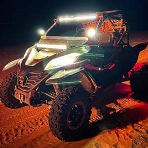 Un buggy de dunas está aparcado en la arena de noche con sus potentes faros y barras de luces encendidos, iluminando el terreno.
