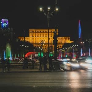 Un imponente edificio illuminato di notte, con fontane colorate e scie luminose a lunga esposizione delle auto su una strada di città.