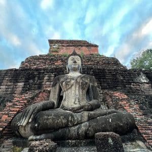Un'imponente statua in pietra del Buddha, segnata dal tempo, siede in posa meditativa di fronte ad antiche rovine in mattoni sotto un cielo nuvoloso.