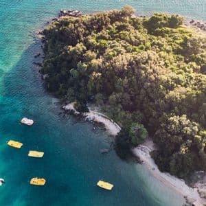 An aerial view of a small, tree-covered island in turquoise waters, with several yellow pedal boats floating nearby.