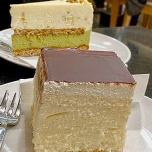 Dos rebanadas de pastel en platos blancos, con un pastel de mousse cuadrado glaseado de chocolate en primer plano y un pastel de pistacho en capas en el fondo.