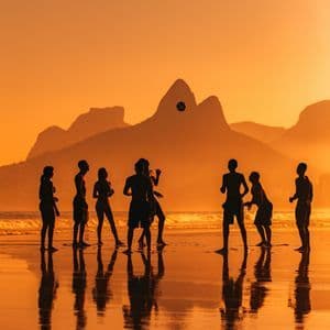 Un viaggio di gruppo WeRoad con persone in silhouette che giocano a palla su una spiaggia bagnata al tramonto, con montagne sullo sfondo.
