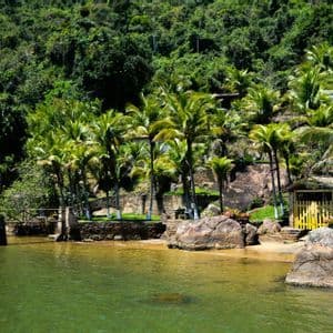 Panorama di un'insenatura tropicale con piccola spiaggia sabbiosa, palme e una capanna, sullo sfondo una lussureggiante collina boscosa.