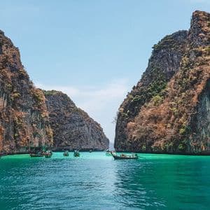 Long-tail boats float on turquoise water between two large, rocky cliffs covered in trees under a clear sky.