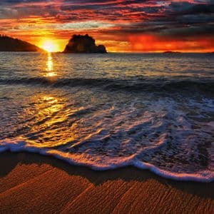Un'onda delicata con schiuma bianca accarezza una spiaggia sabbiosa durante un tramonto colorato sull'oceano, riflettendosi sull'acqua.
