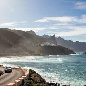 Un'auto percorre una strada tortuosa lungo una costa frastagliata, con montagne lontane e un piccolo villaggio sotto un cielo soleggiato.