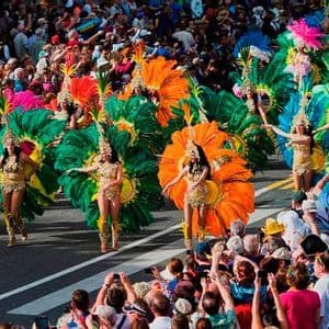 Danzatori in costumi colorati e piumati sfilano per una strada, osservati da una grande folla di spettatori.