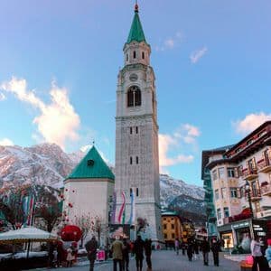 Un alto campanile in pietra con una guglia verde si staglia su una piazza animata da persone, con lo sfondo di montagne innevate.