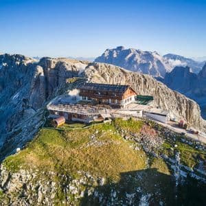 Un rifugio di montagna con un'ampia terrazza si trova su una cima erbosa, circondato da vette rocciose sotto un cielo azzurro e limpido.