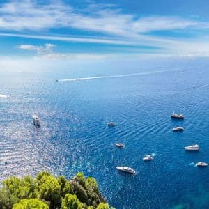 Una vista panoramica di una costa rocciosa con un grande faraglione, che si affaccia su un mare azzurro brillante punteggiato da numerose barche e yacht in una giornata di sole.
