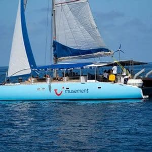 Un gran catamarán azul claro con pasajeros a bordo navega en mar abierto con sus velas desplegadas.
