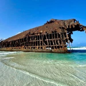 Un gran naufragio oxidado descansa en una playa de arena, parcialmente sumergido en aguas cristalinas de color turquesa bajo un cielo azul.