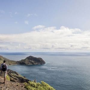 Un escursionista con uno zaino si trova su un sentiero costiero roccioso con vista sull'oceano e un promontorio distante sotto un cielo parzialmente nuvoloso.