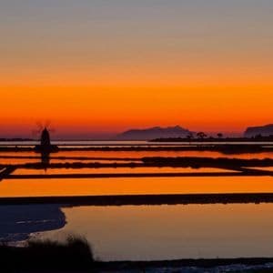 Una sagoma di mulino a vento si staglia sulle saline che riflettono un vibrante tramonto arancione, con le montagne in lontananza.