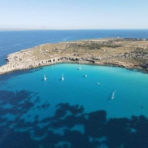 Una vista aerea di barche a vela in una baia turchese accanto a una penisola rocciosa sotto un cielo sereno.