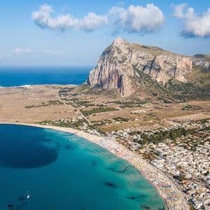 Veduta aerea di una cittadina costiera e di una spiaggia affollata, lambite da acque turchesi, con una grande montagna rocciosa sullo sfondo.
