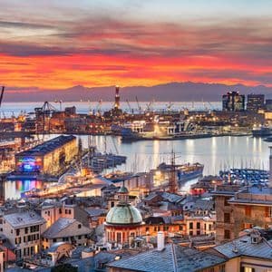 Una vista panoramica di una grande città portuale al tramonto, con edifici illuminati e barche nel porto sotto un cielo colorato.