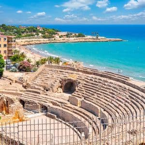 Un antico anfiteatro in pietra si affaccia su una spiaggia sabbiosa con acqua turchese e una città costiera sotto un cielo parzialmente nuvoloso.