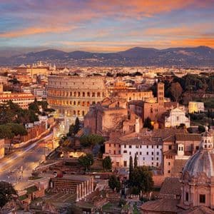 Veduta aerea del Colosseo e del panorama urbano circostante al tramonto, con montagne all'orizzonte sotto un cielo colorato.